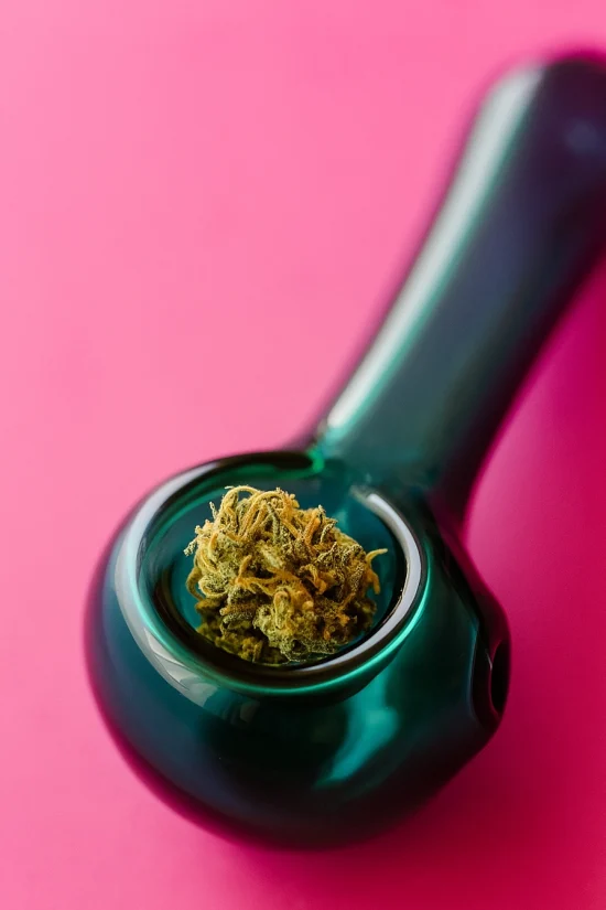 Close-up of a teal glass weed pipe filled with cannabis on a pink background.